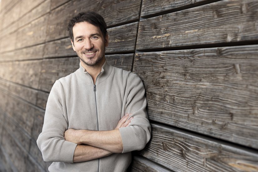 Das Bild zeigt einen Mann mittleren Alters mit braunen Haaren und einem drei Tage Bart, in beigefarbenen Pullover und verschränkten Armen. Er lehnt lächelnd an einer Holzwand.